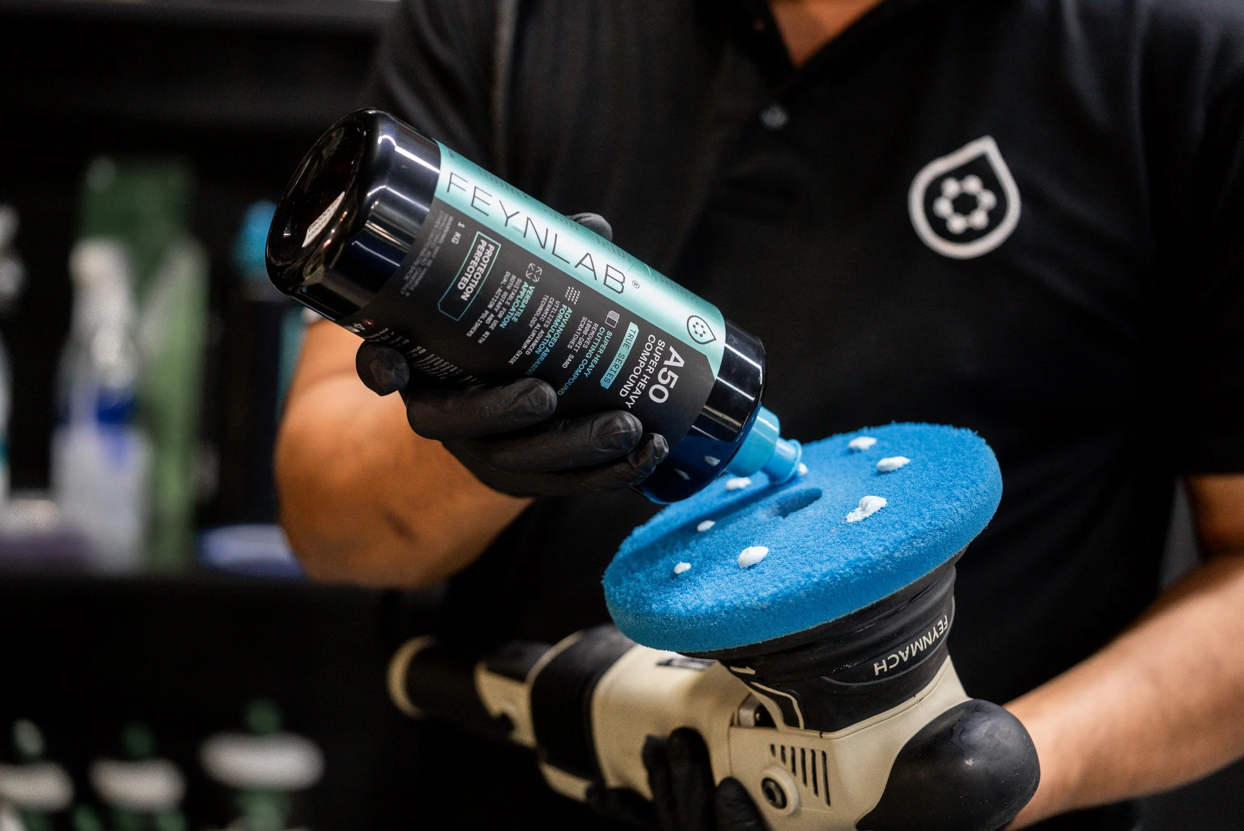 A person using a power buffer with a blue polishing pad, applying a compound from a bottle labeled 'FEYNLAB' to polish a surface.