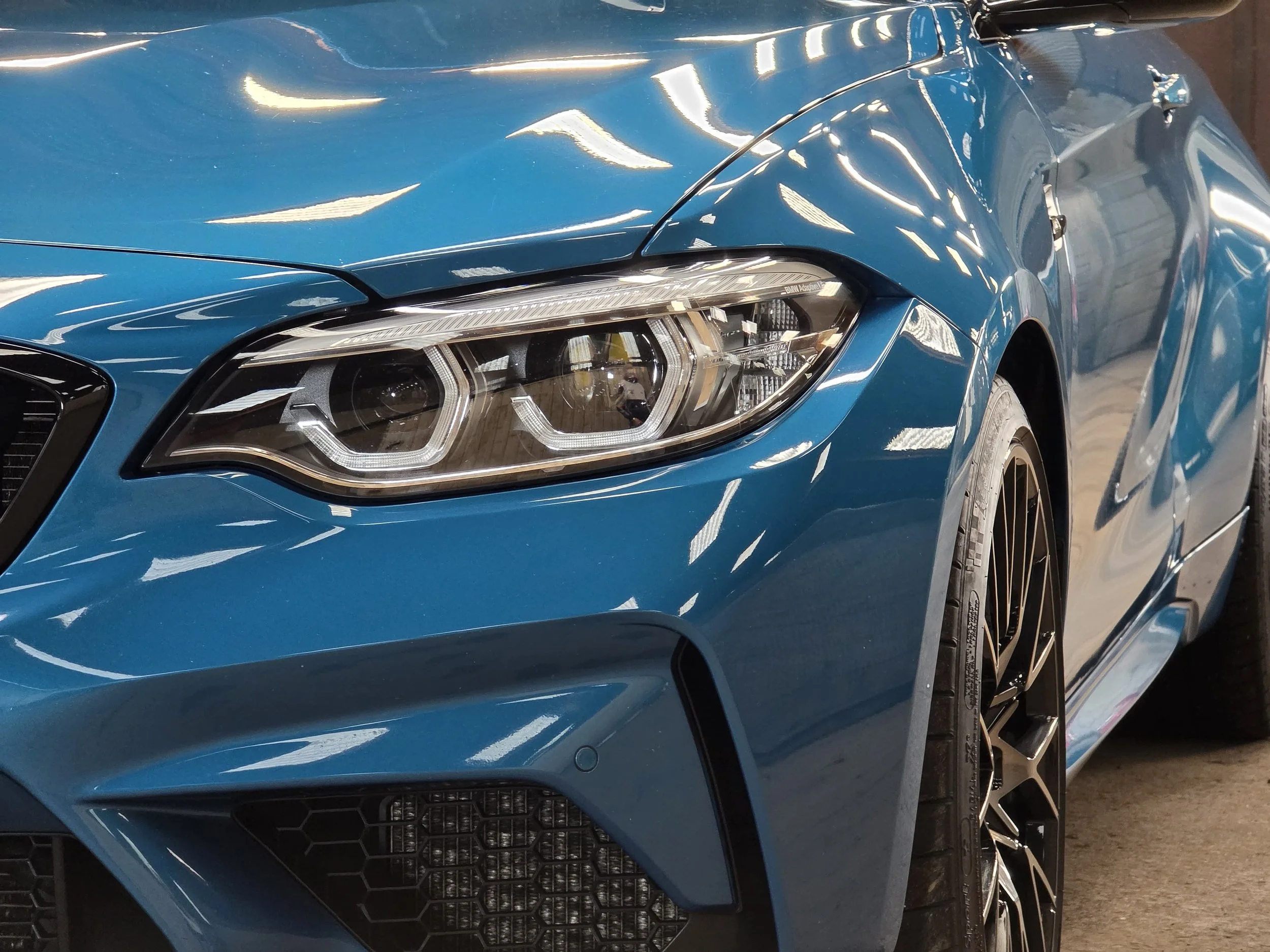Close-up of a blue luxury car's front left side, showing the headlight, wheel, and part of the hood.