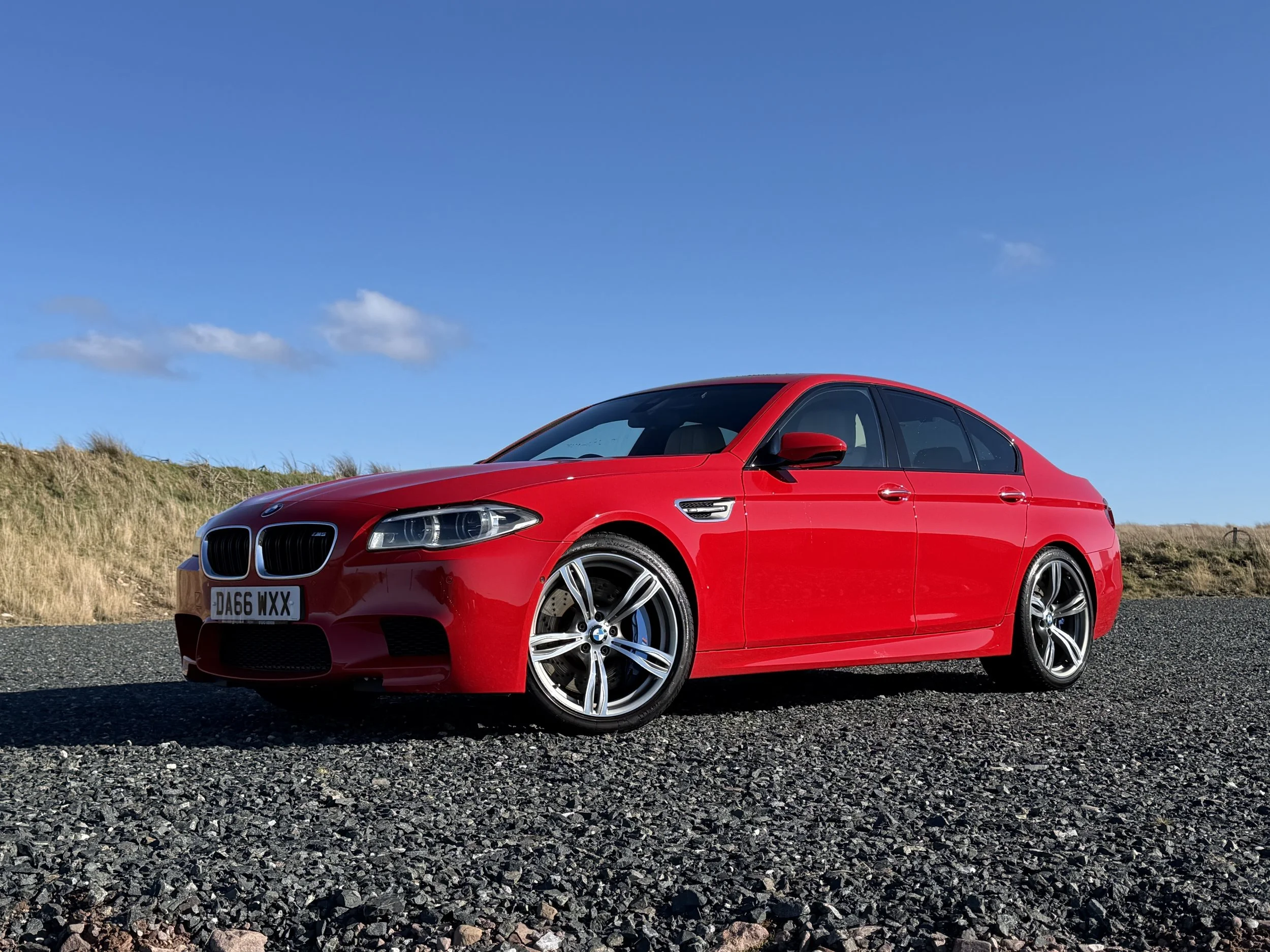 A red BMW sedan parked on a gravel surface under a blue sky with a few clouds and grassy hillside in the background.