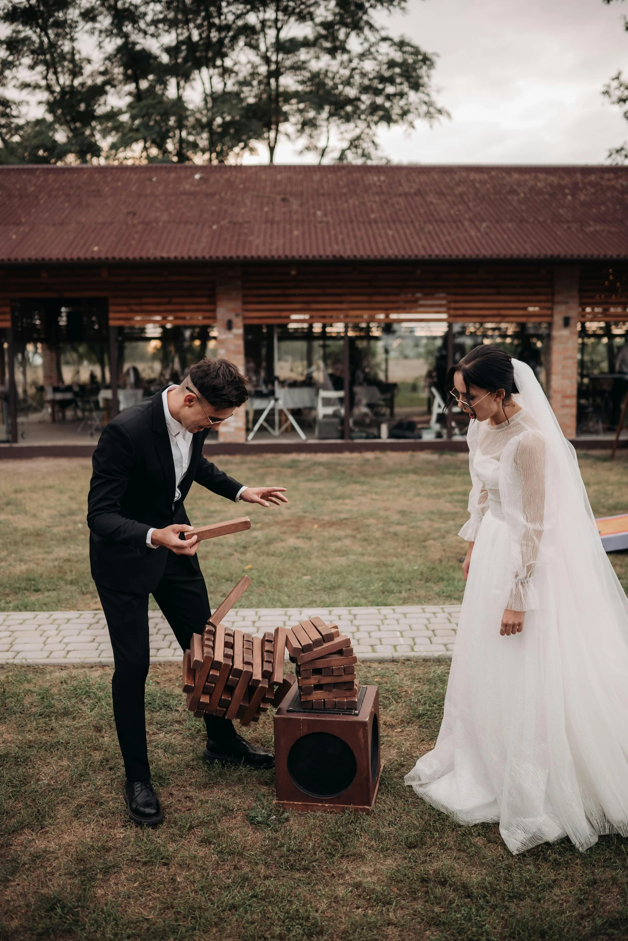 A bride in a white wedding dress laughing while a groom in a black suit plays a game of Jenga outdoors at a wedding reception.