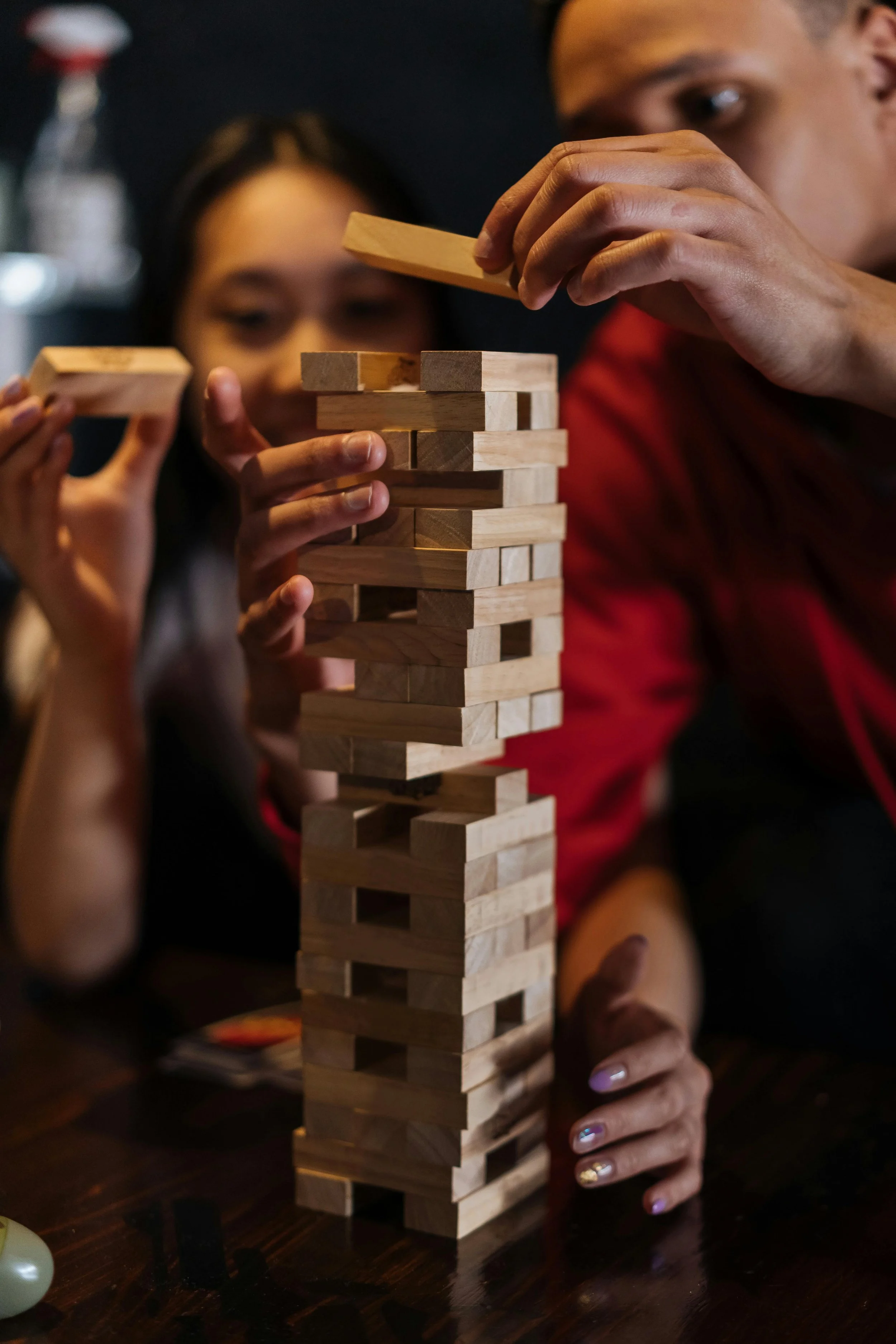 People playing a game of Jenga, removing a piece from a wooden block tower in a dimly lit setting.
