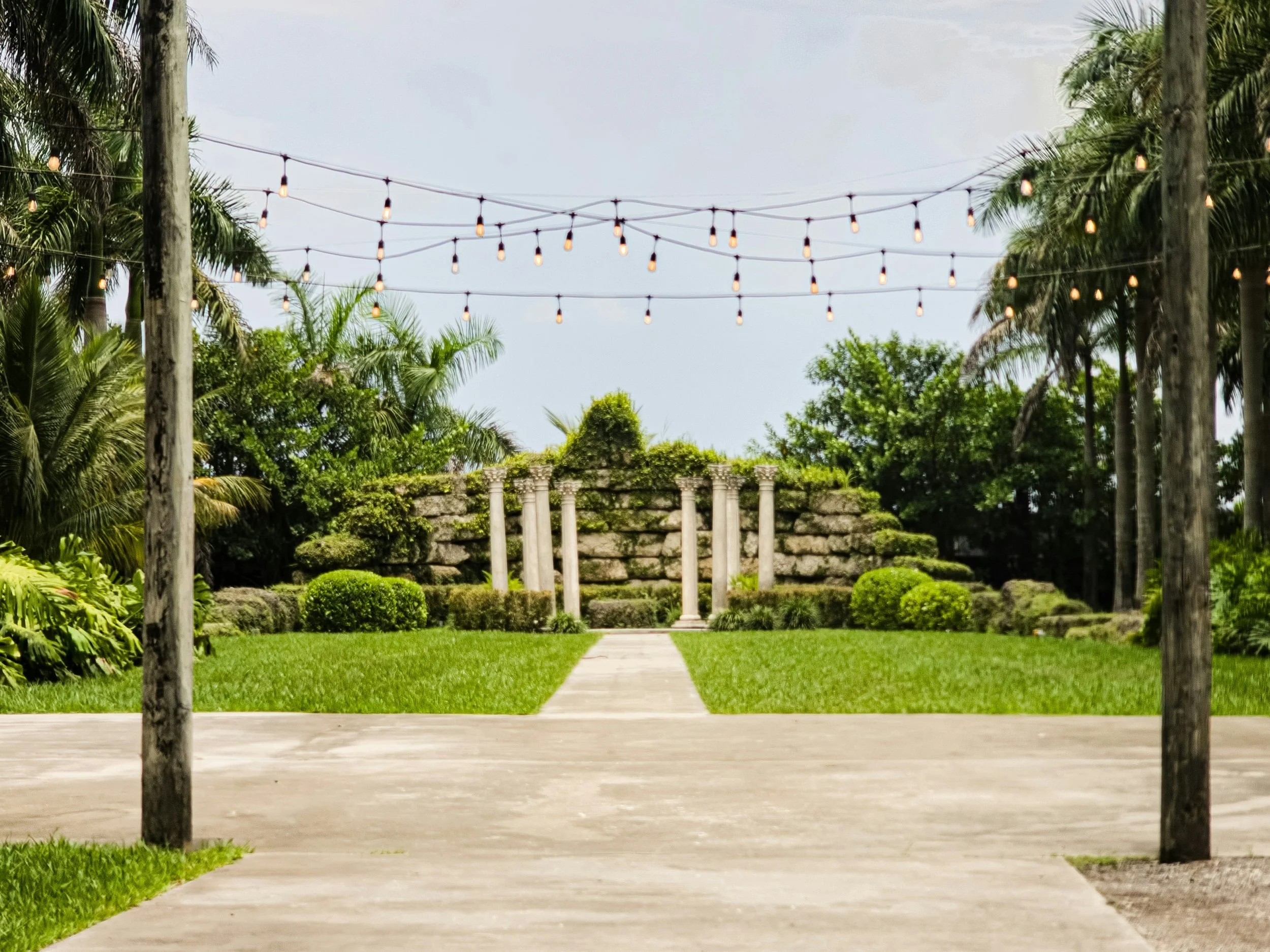 Outdoor garden with stone steps, columns, green bushes, palm trees, and string lights overhead.