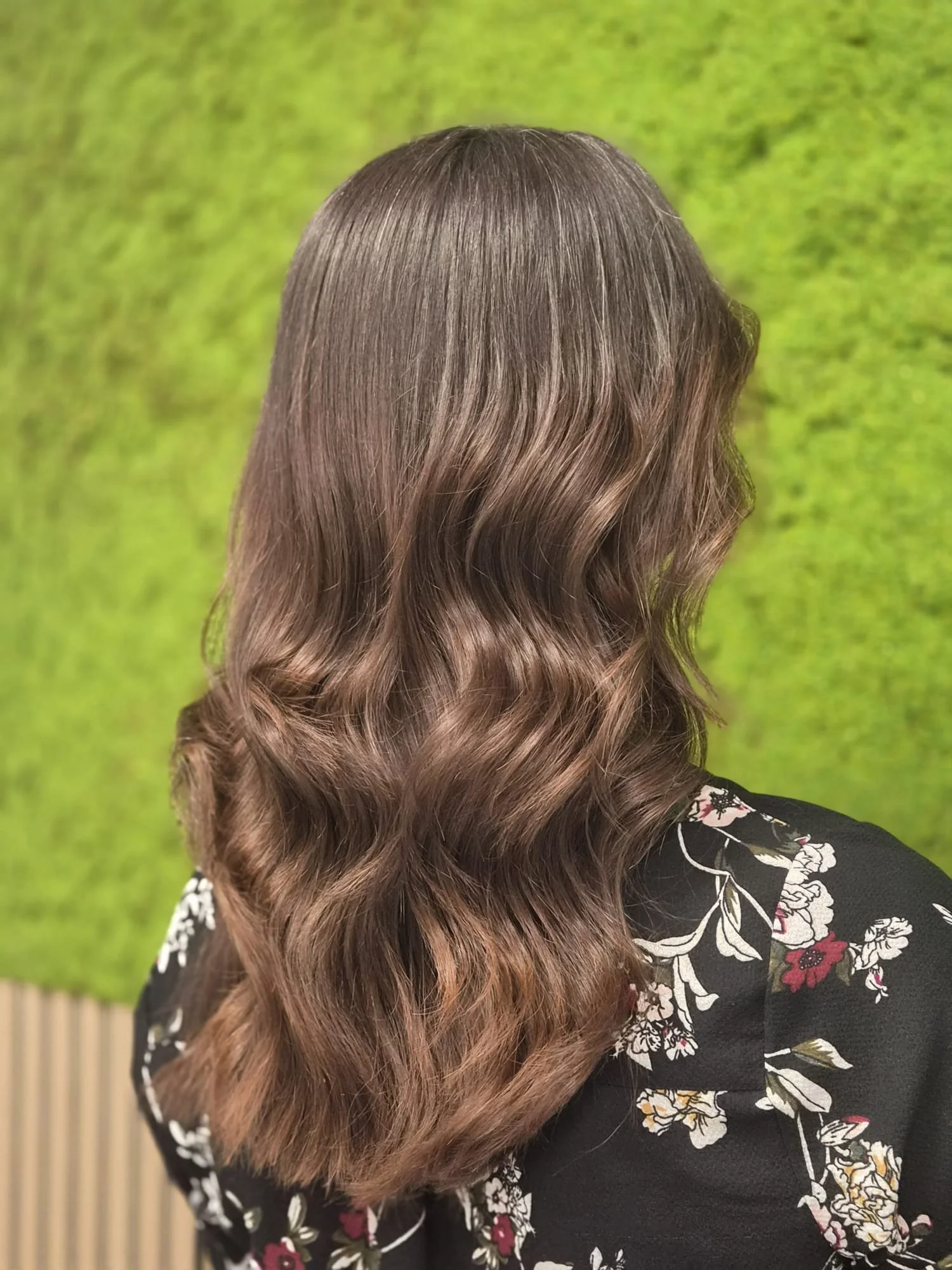 Frontalaufnahme einer Person mit langen, glatten braunen Haaren und einer schwarzen Bluse mit weißen und roten Blumenmuster. Hintergrund grüne Wände.