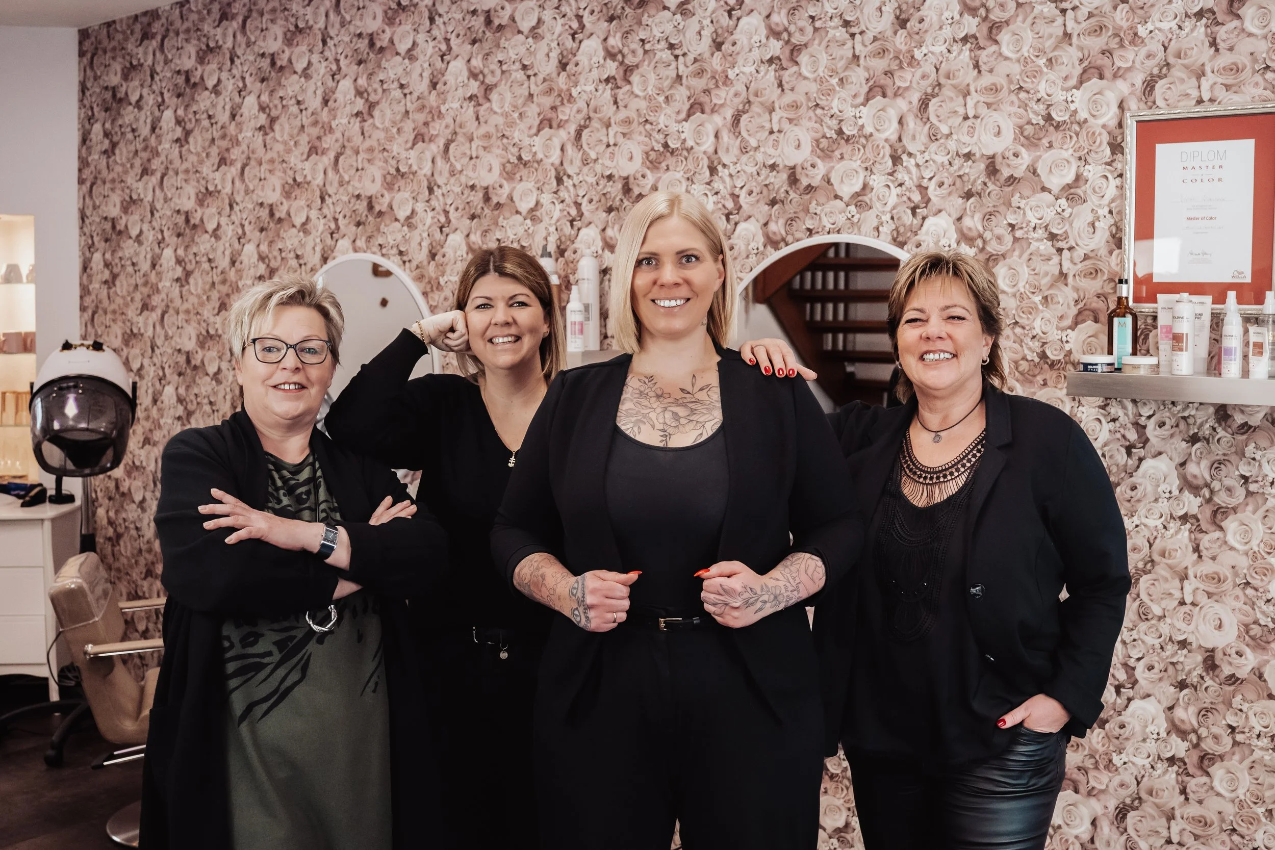Vier Frauen stehen vor einer rosafarbenen Rosenwand im Innenraum, lächeln in die Kamera, einige haben Tattoos und tragen schwarze Kleidung.