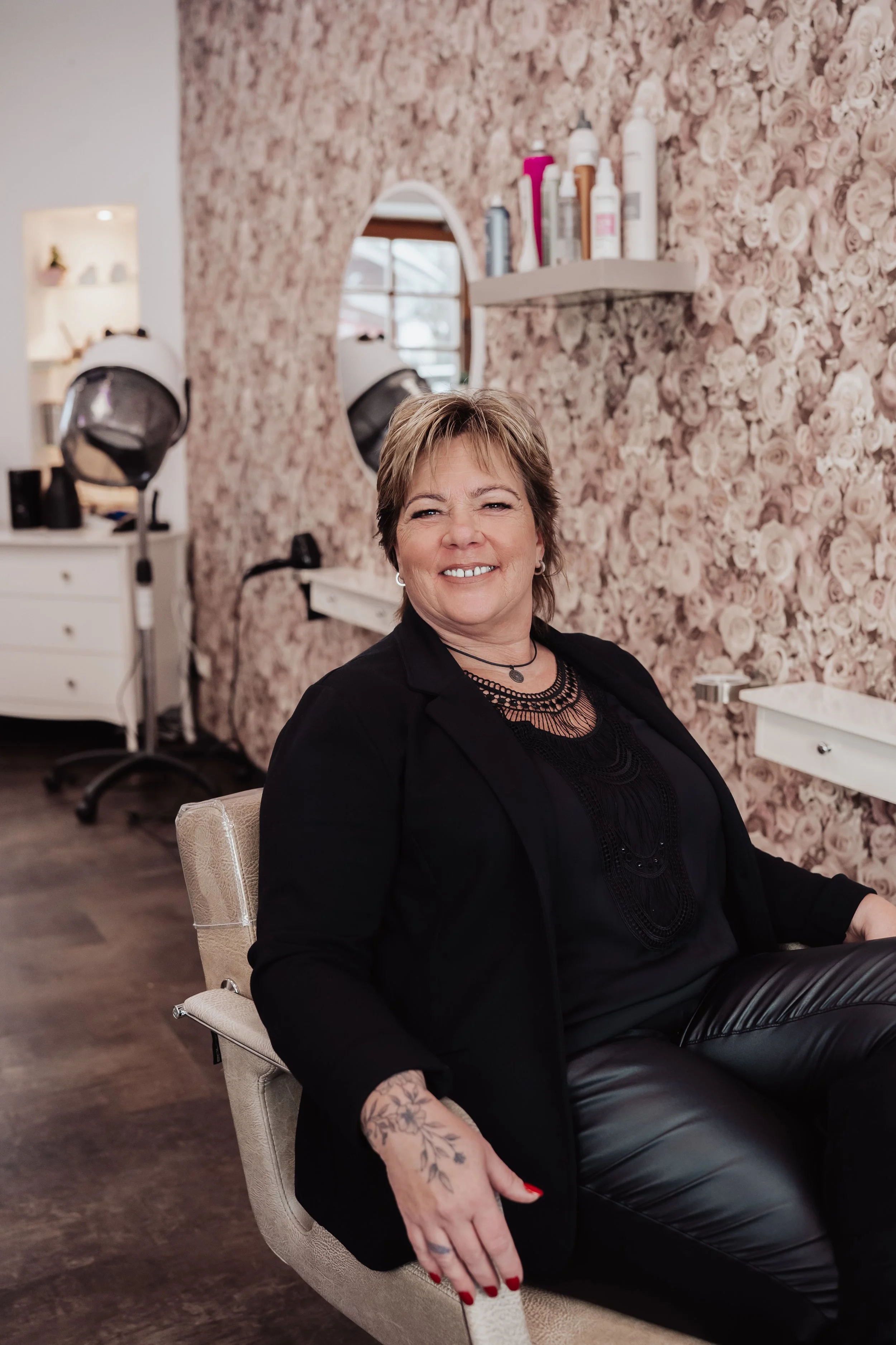 Eine lächelnde Frau sitzt in einem stylischen Friseursalon vor einer blumigen Wandtapete, mit Haartrocknern im Hintergrund und einem Spiegel. Sie trägt schwarzes Kleidung und Halsketten, hat kurze braune Haare, Tattoos auf ihrer Hand, rote Lippen und Nägel.