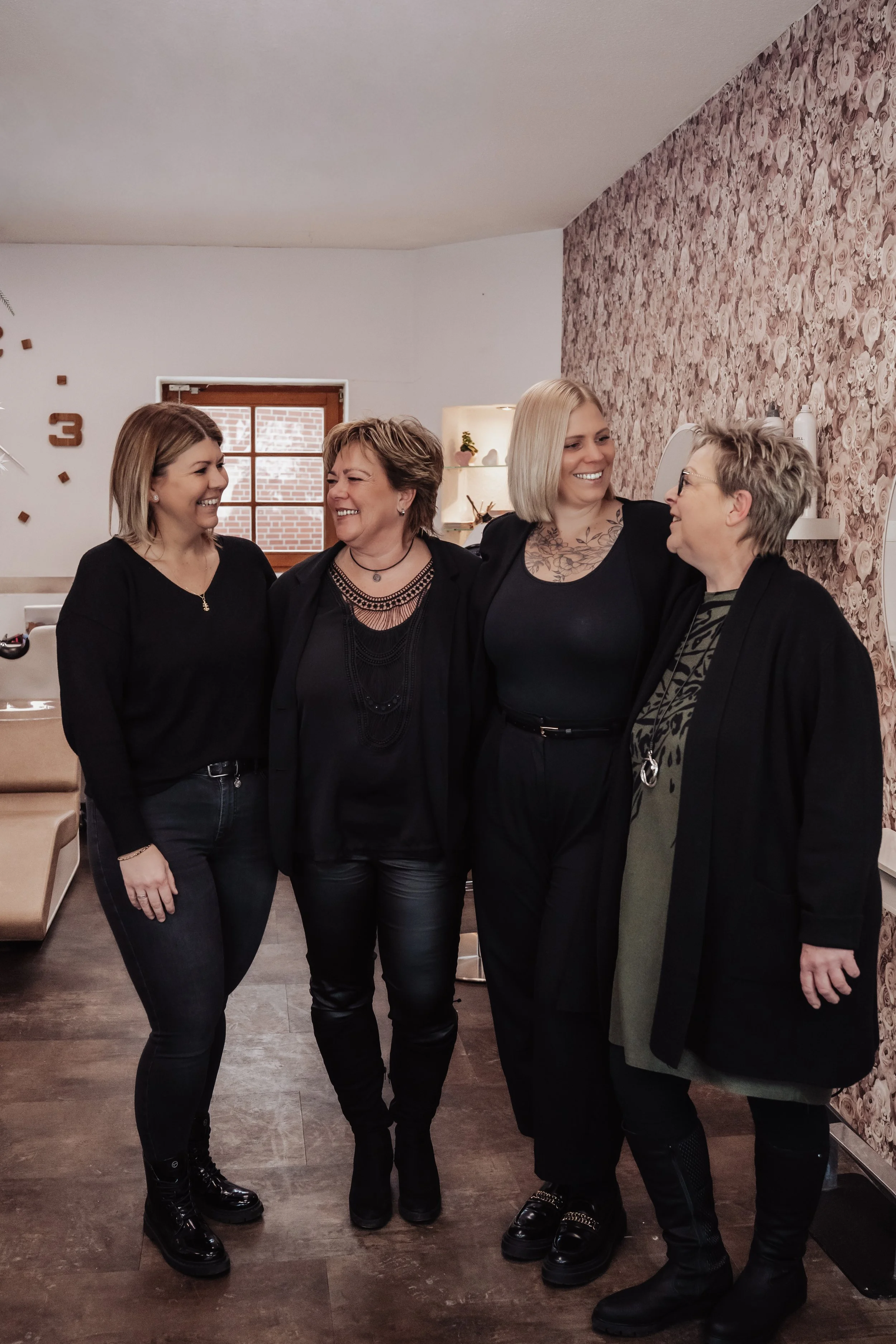 Fünf Frauen, die sich umarmen und lachen, in einem Raum mit floral an der Wand dekoriert und Holzfußboden, die meisten tragen dunkle Kleidung.