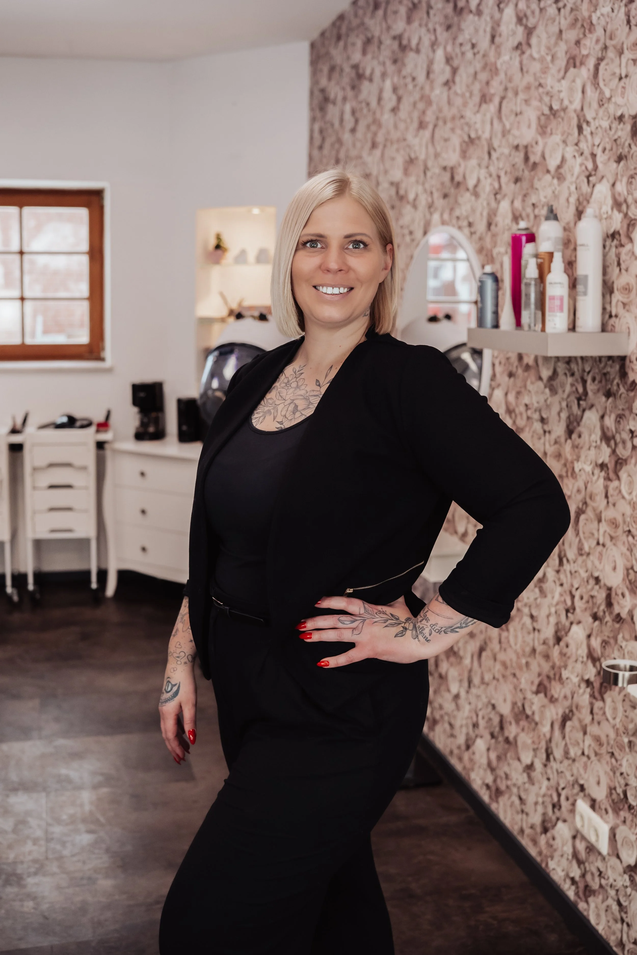 Eine Frau mit kurzen blondem Haar, Tattoos auf Armen und Dekolleté, trägt schwarze Kleidung und pose in einem Raum mit floral gekachelter Wand im Hintergrund.