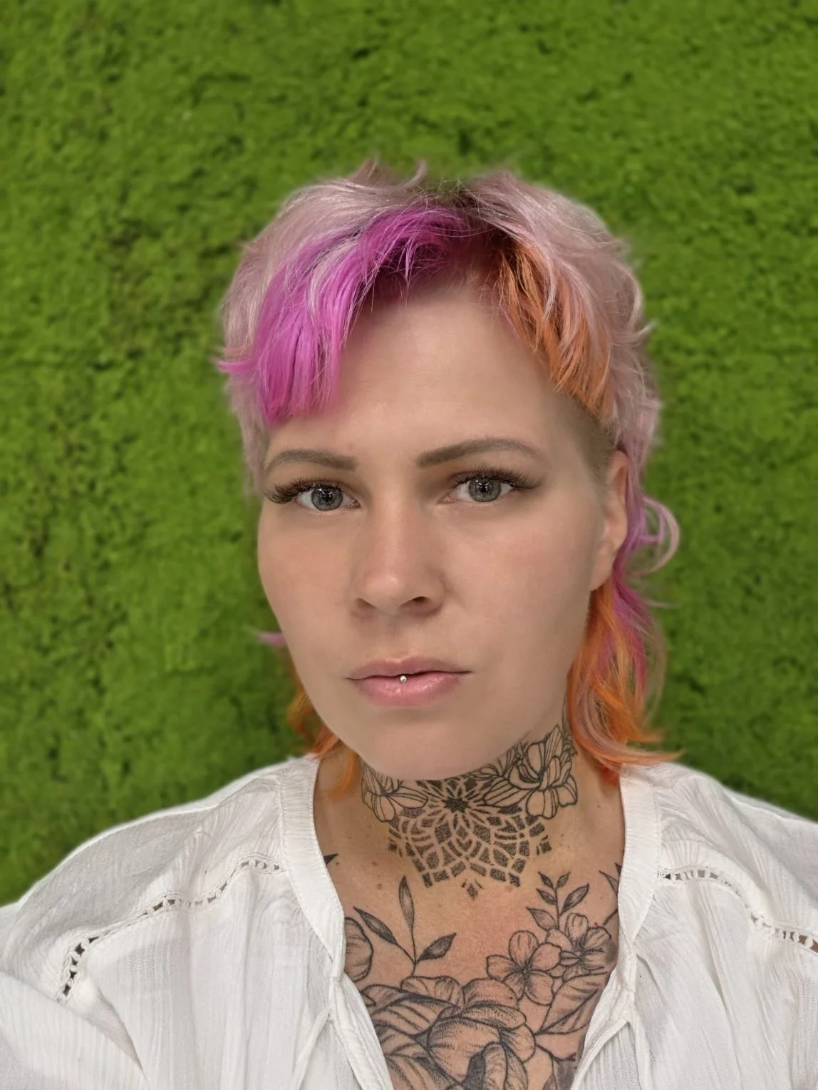 Junge Frau mit pink und oranges Haar, Tätowierungen im Dekolleté, Piercing unter der Oberlippe, steht vor grünem Hintergrund und blickt in die Kamera.