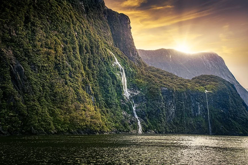 Waterfalls flowing down cliffs at Milford Sound with golden sunset light over the fiord.