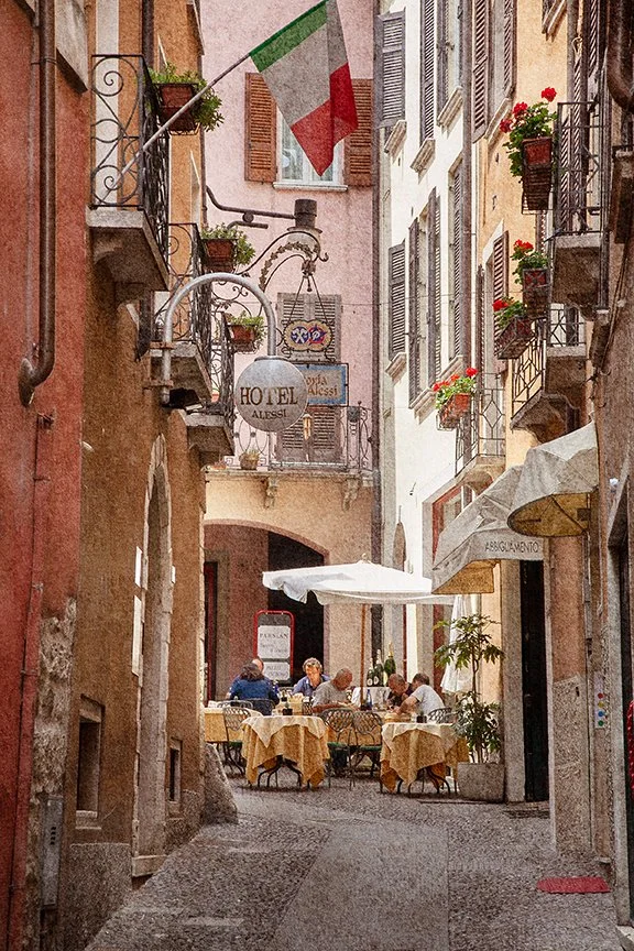 Italian village laneway with café diners, balconies, flowers and Italian flag.