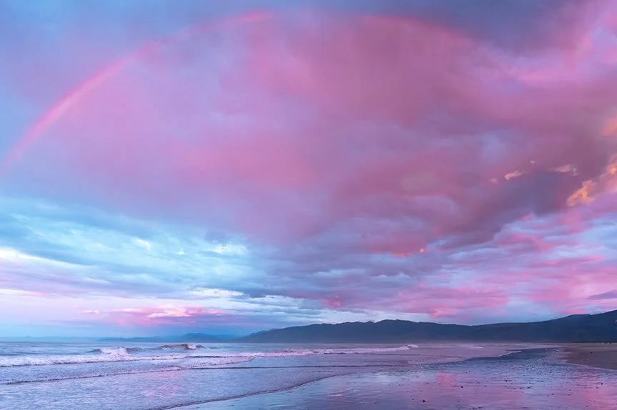 Rainbow arching across a pastel sunset sky above the ocean shoreline.