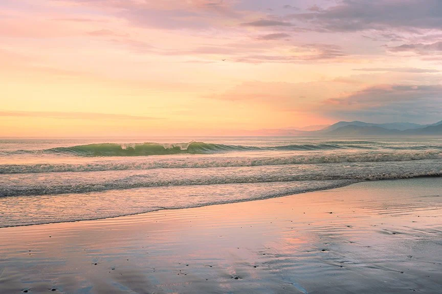 Soft ocean waves at sunrise with pastel sky and distant mountains.