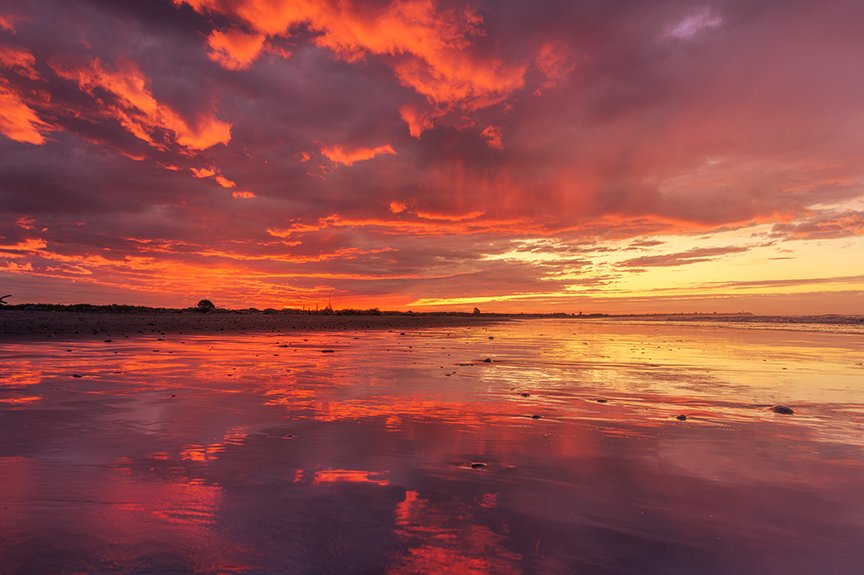 Dramatic red and orange sunset reflecting across wet beach sand.
