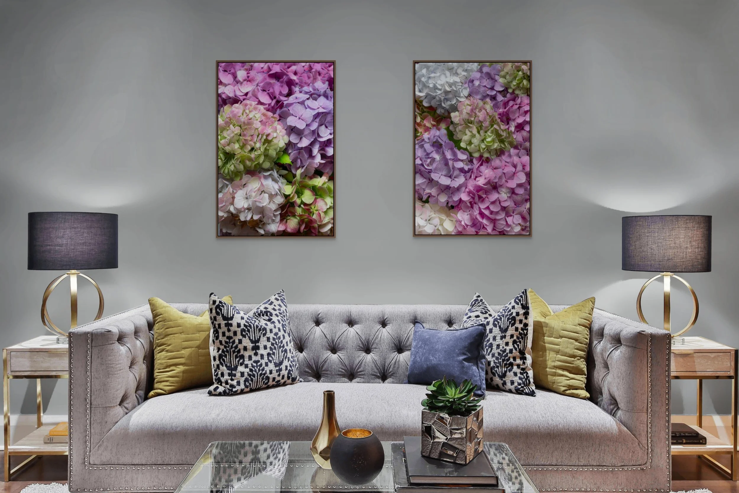 Living room with a gray tufted sofa decorated with various yellow and patterned throw pillows, a glass coffee table with decorative objects, two side tables with black lamps, and two framed floral artwork pieces on a light gray wall.