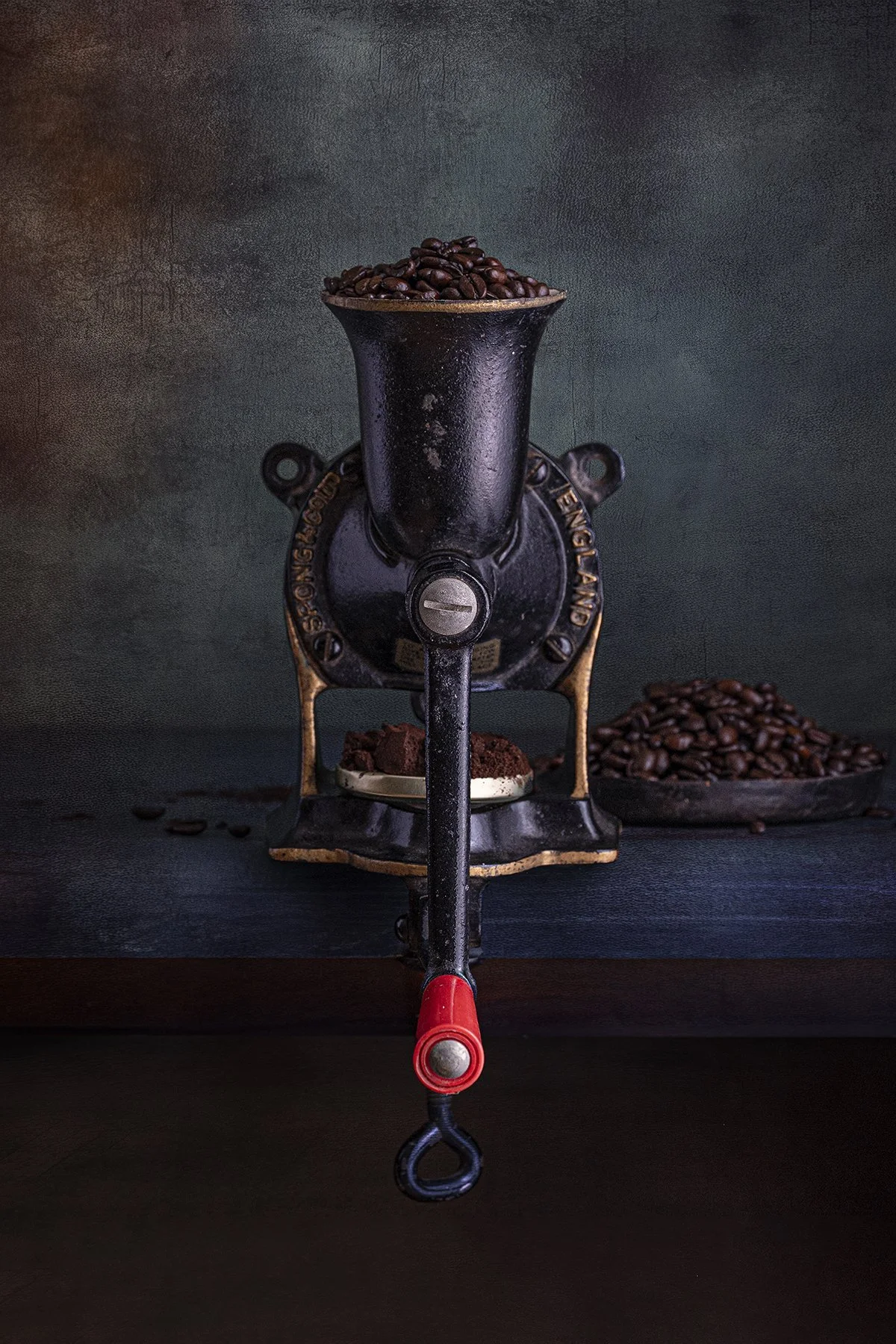 Still life fine art photograph of a vintage hand-crank coffee grinder stands proudly against a richly textured backdrop, filled with freshly roasted, by From The Field.