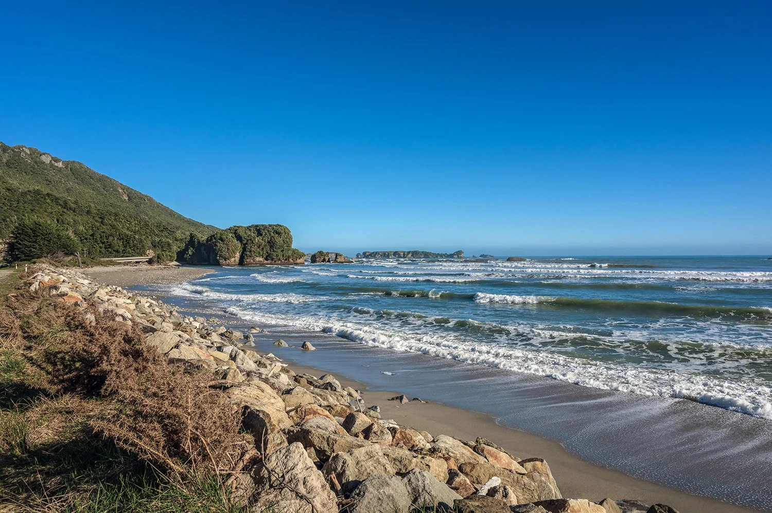 Waves rolling onto a rocky West Coast beach with distant sea stacks beneath a clear blue sky.