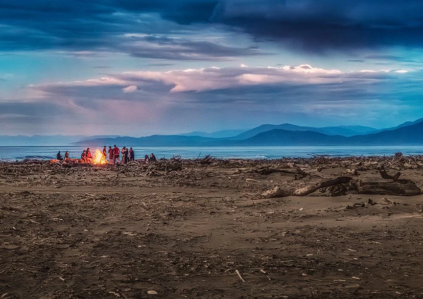 People gathered around a beach bonfire on a driftwood covered coastline at dusk.
