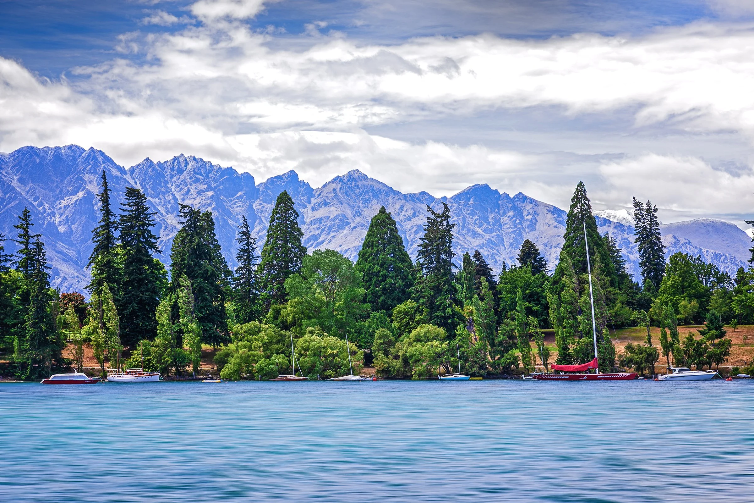 A scenic view of a lake with boats anchored near the shoreline, lush green trees, and majestic snow-capped mountains under a partly cloudy sky.