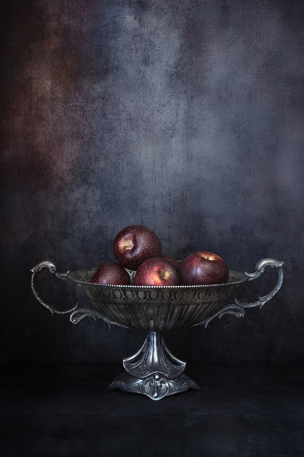 Red apples displayed in an ornate silver bowl against a dark background.