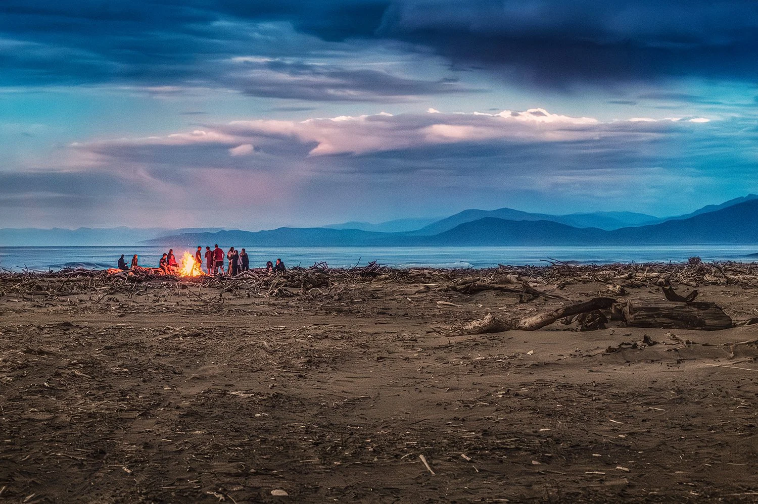 People gathered around a beach bonfire on a driftwood-covered West Coast beach.