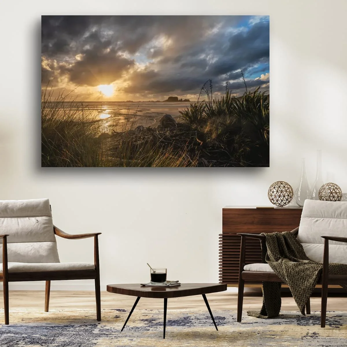 Sun rays breaking through dark coastal clouds over a beach framed by flax, grasses and driftwood.
