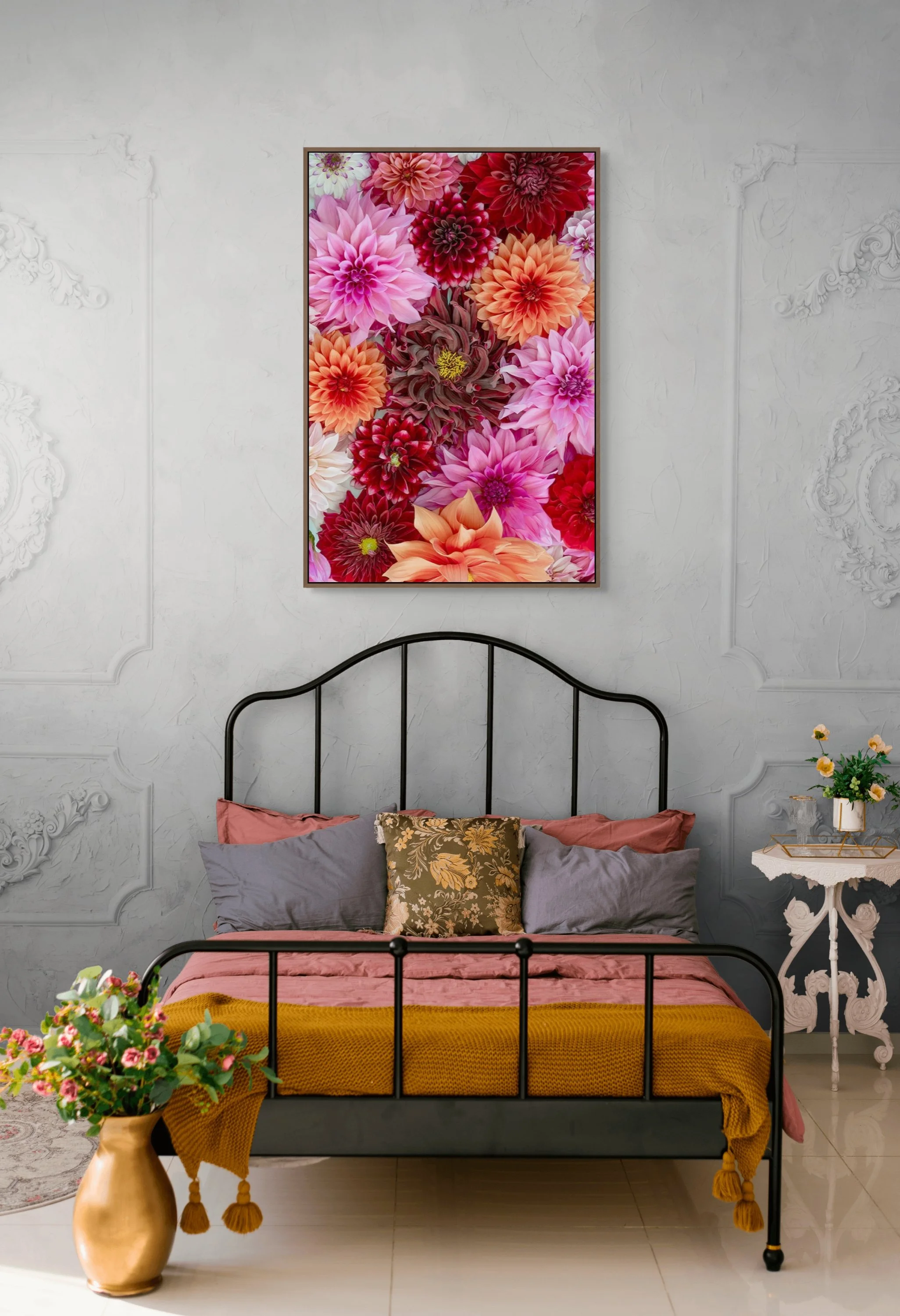A bedroom with a black metal bed frame, pink and gray bedding, and an assortment of pillows. A white ornate side table with a plant in a gold pot is beside the bed. A large floral painting hangs on the wall above, and flowers in a gold vase are on the floor in front of the bed.