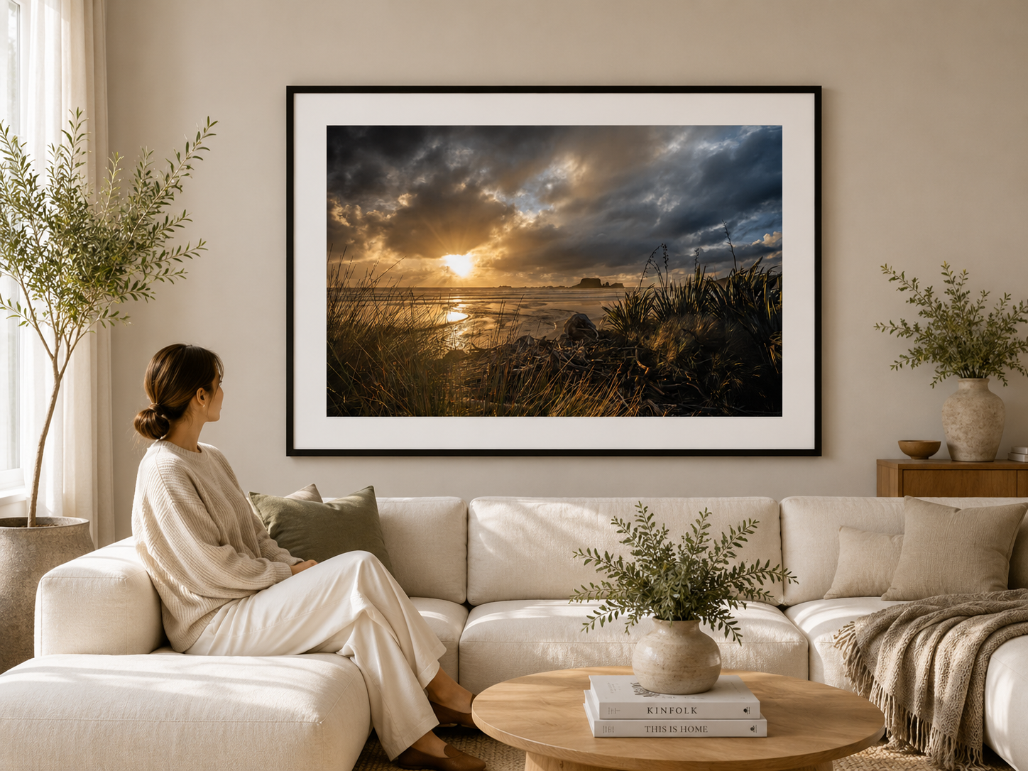 Sun rays breaking through dark coastal clouds over a beach framed by flax, grasses and driftwood.