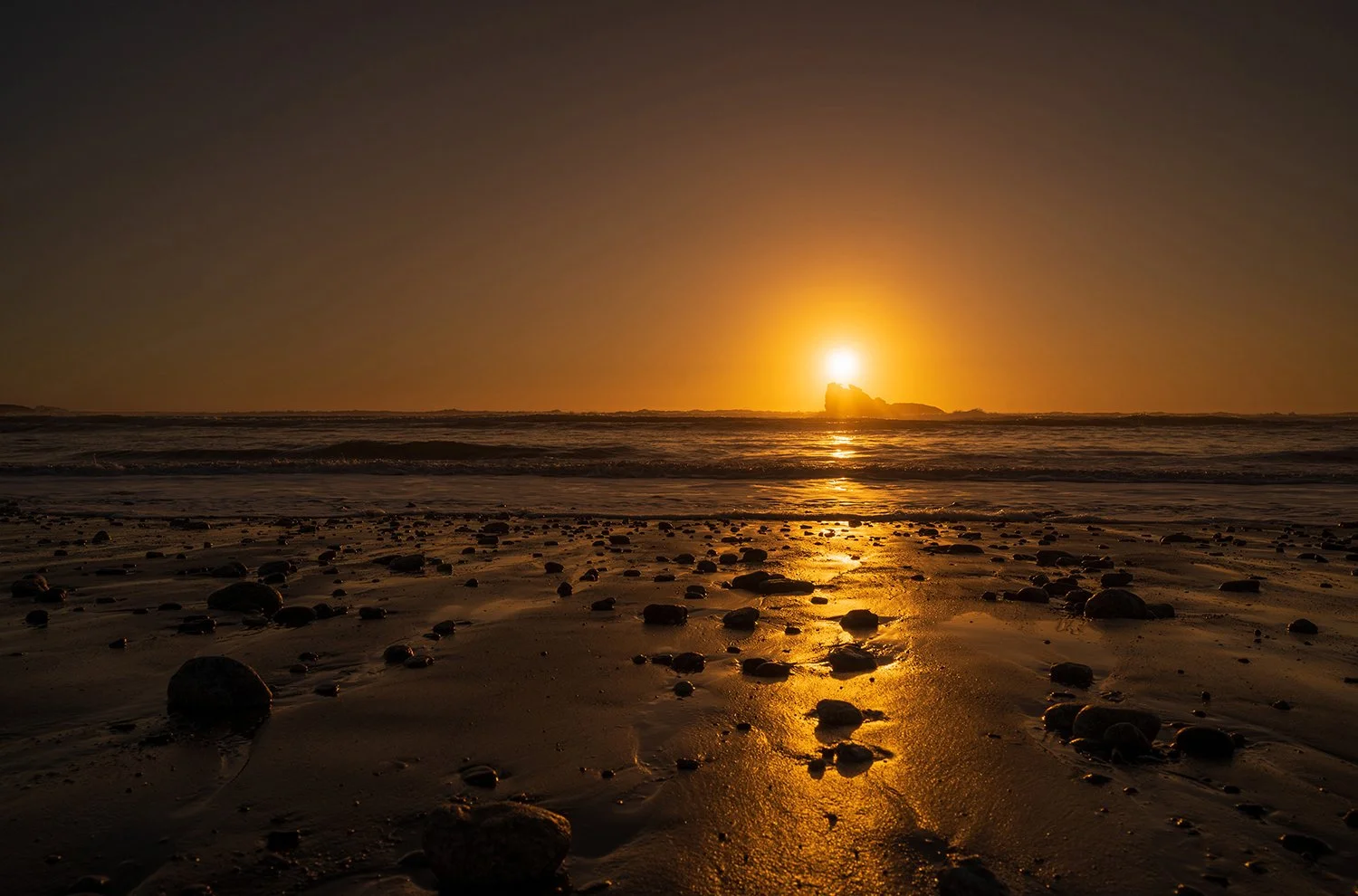 Golden sunset reflecting across wet sand and stones with offshore rock on the West Coast.