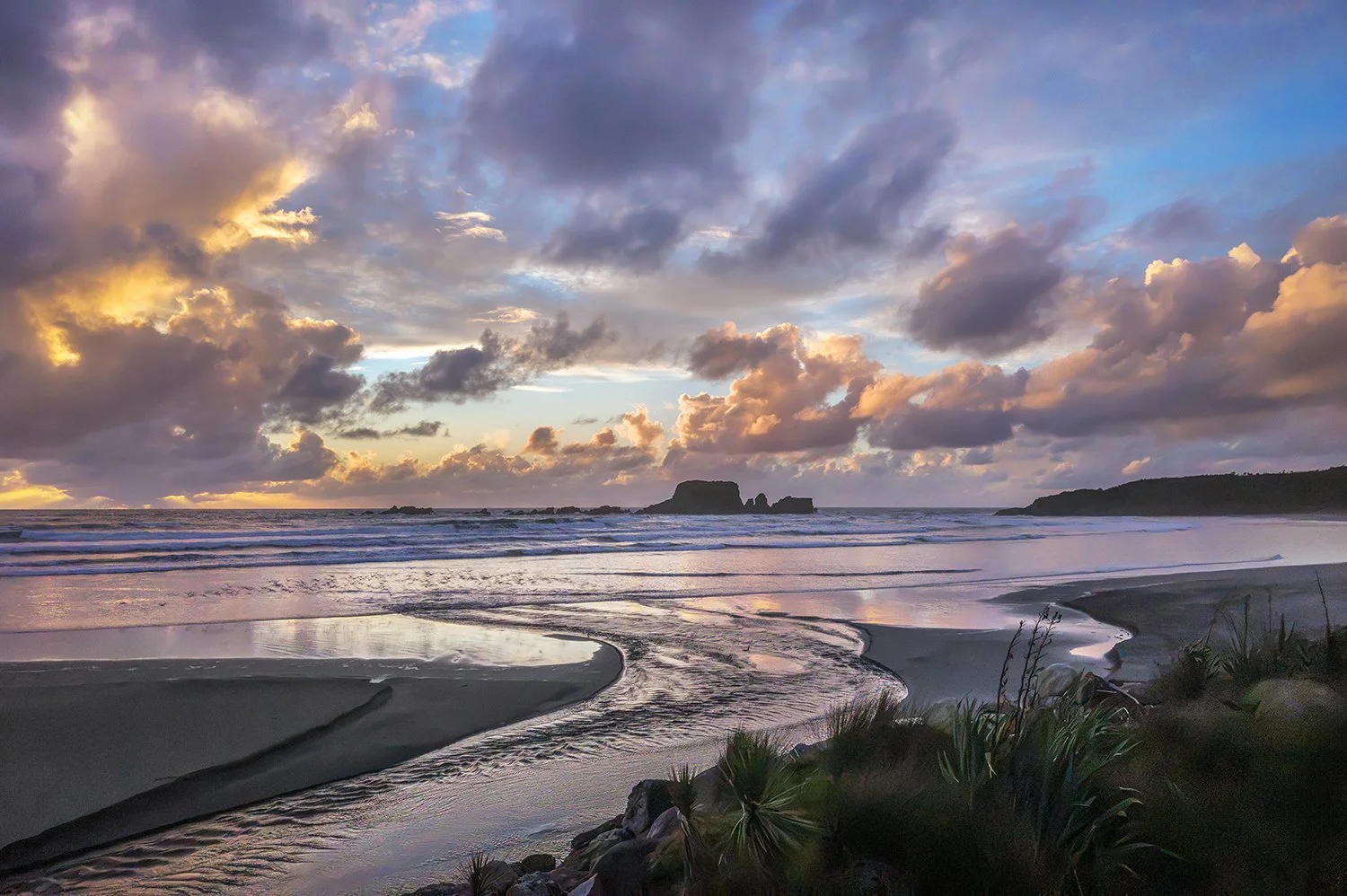 Curving tidal stream on a beach beneath pastel blue, mauve and peach clouds with offshore rock formations.
