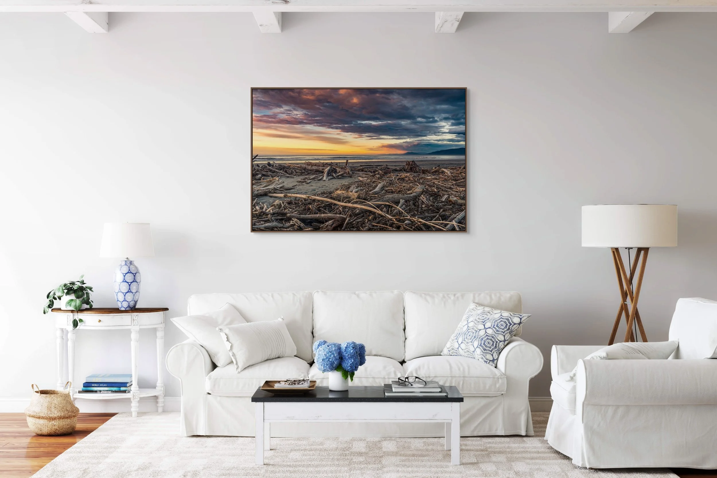 Interior of a living room with a white sofa, a coffee table, and decor including a blue and white vase, books, and pillows. A large framed sunset or sunrise photograph hangs on the wall. There are matching lamps and a wicker basket on the floor.