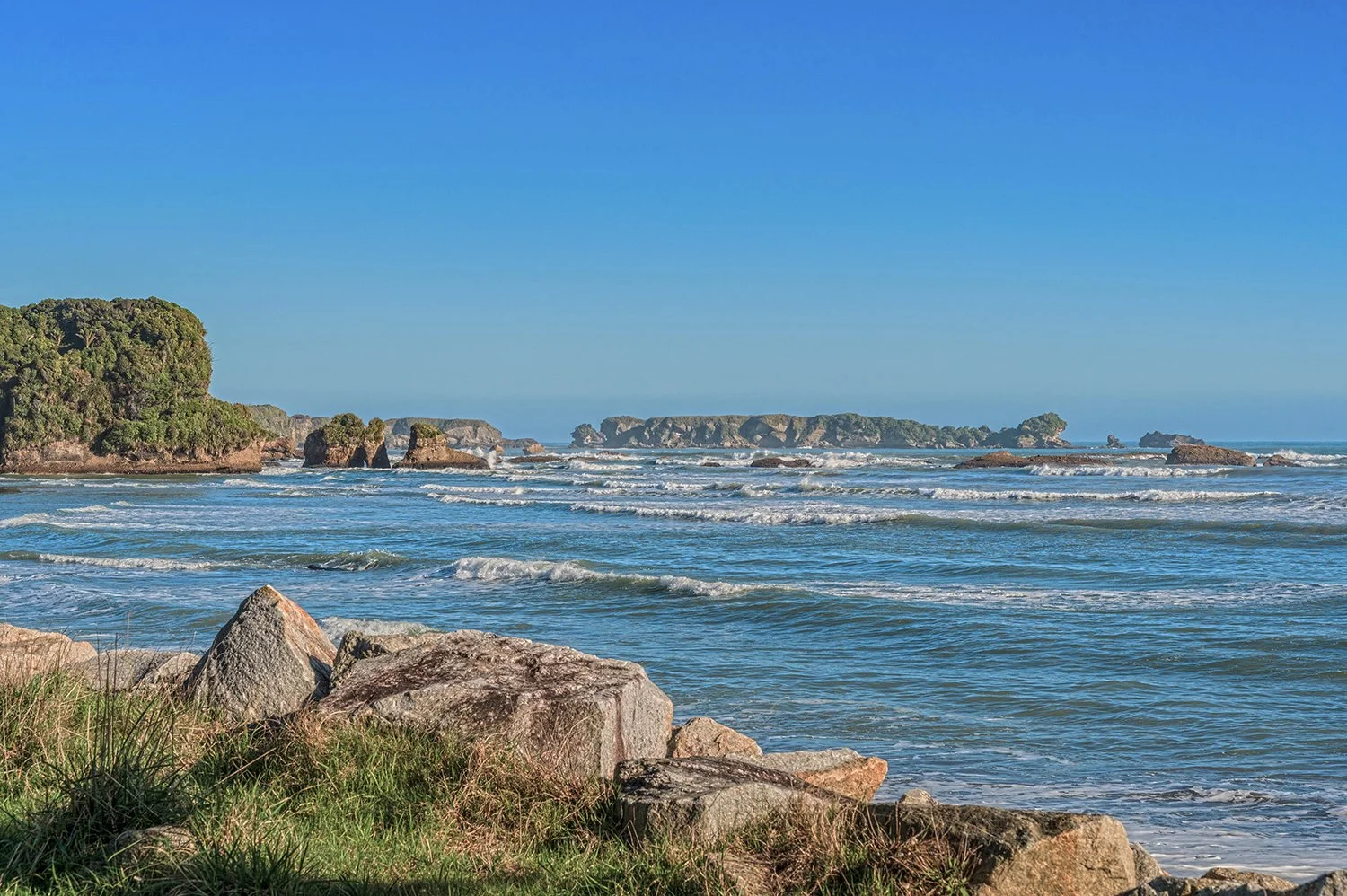 Waves rolling towards rocky coastline beneath blue skies on the West Coast of New Zealand.
