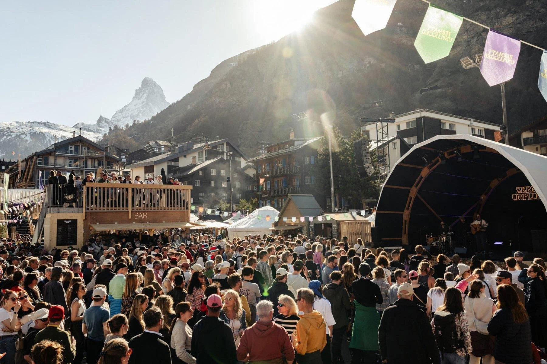 A large outdoor music concert with a crowd of people gathered in front of a stage where musicians are performing. The scene takes place in a mountainous area with houses and trees in the background, under a bright sun.