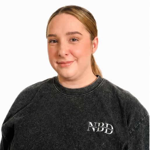 A young woman with light skin and light brown hair tied back, wearing a dark gray sweatshirt with a small logo on the chest that says 'NBD', standing against a plain white background.