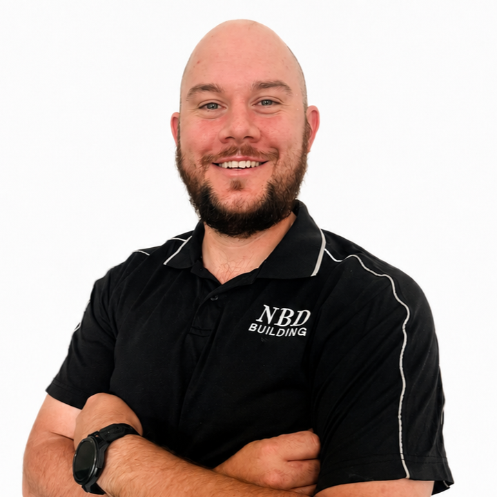 A smiling man with a beard and bald head, wearing a black polo shirt with white piping and an 'NBD Building' logo, standing with arms crossed against a white background.
