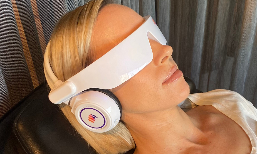 A woman with blonde hair relaxing with a white virtual reality headset and headphones on, lying on a black lounge chair.