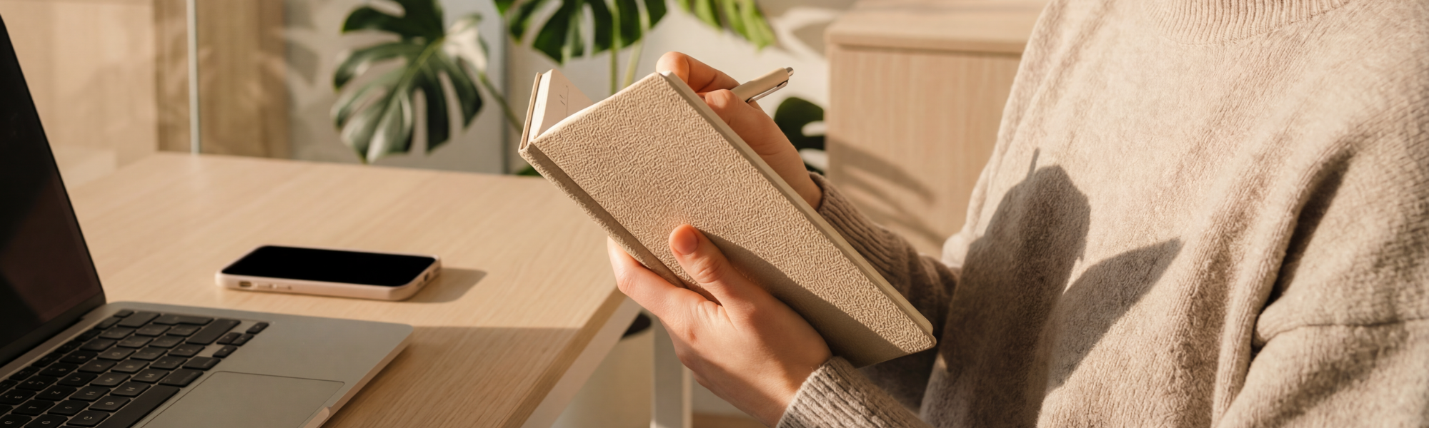 Person holding a beige textured notebook and pen at a desk with a laptop and smartphone, with a potted plant in the background.