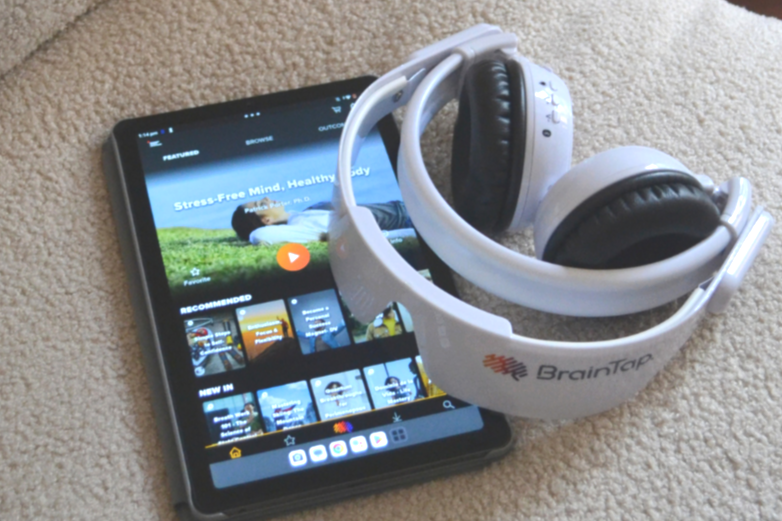 A smartphone displaying a meditation app next to a white BrainTap headset on a beige surface.