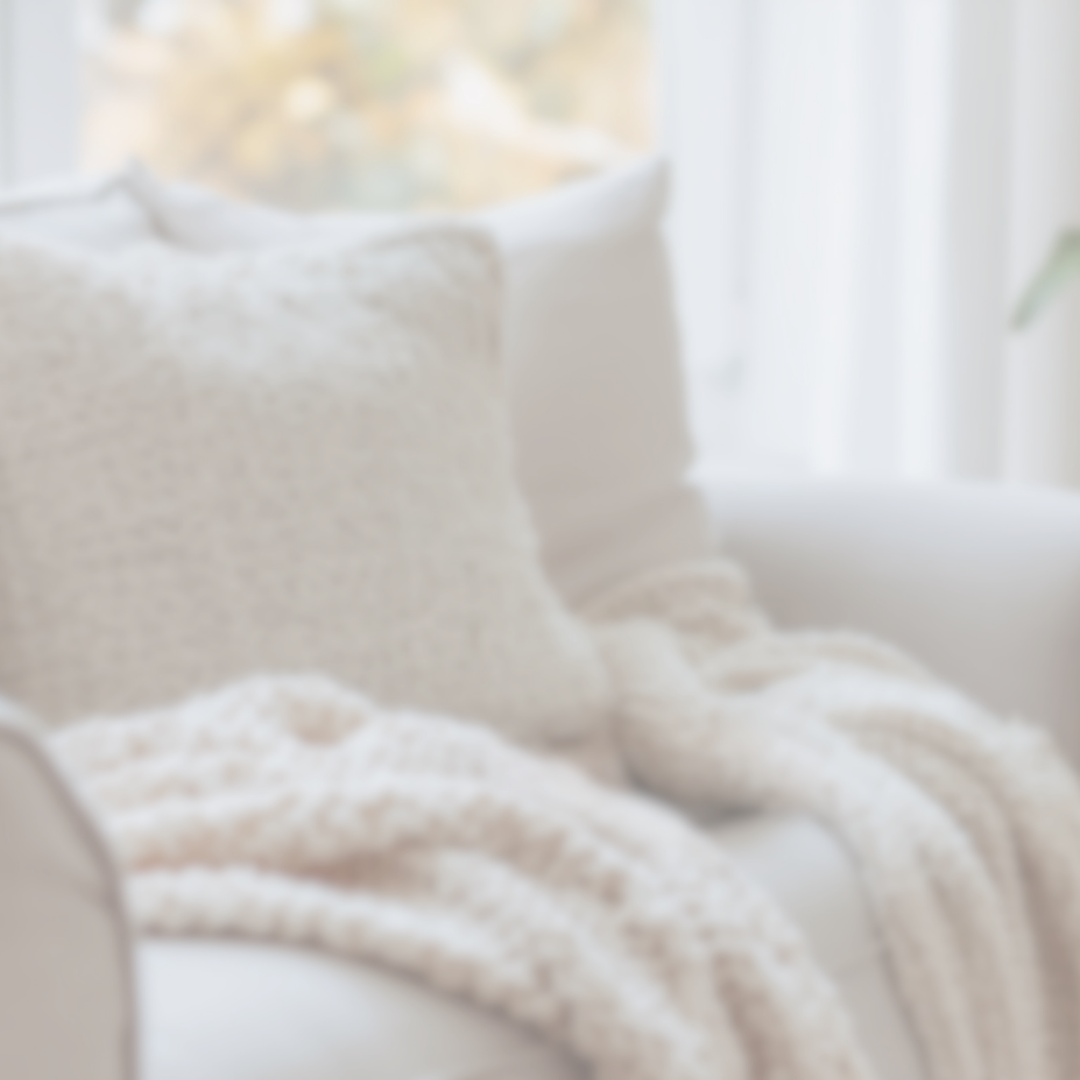 A soft, cushioned sofa with cream-colored pillows and a cozy throw blanket near a window with white curtains.