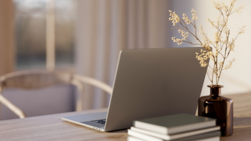 A silver laptop on a wooden desk next to a small brown vase with dried white flowers and stacked books, with sunlight coming through window curtains in the background.