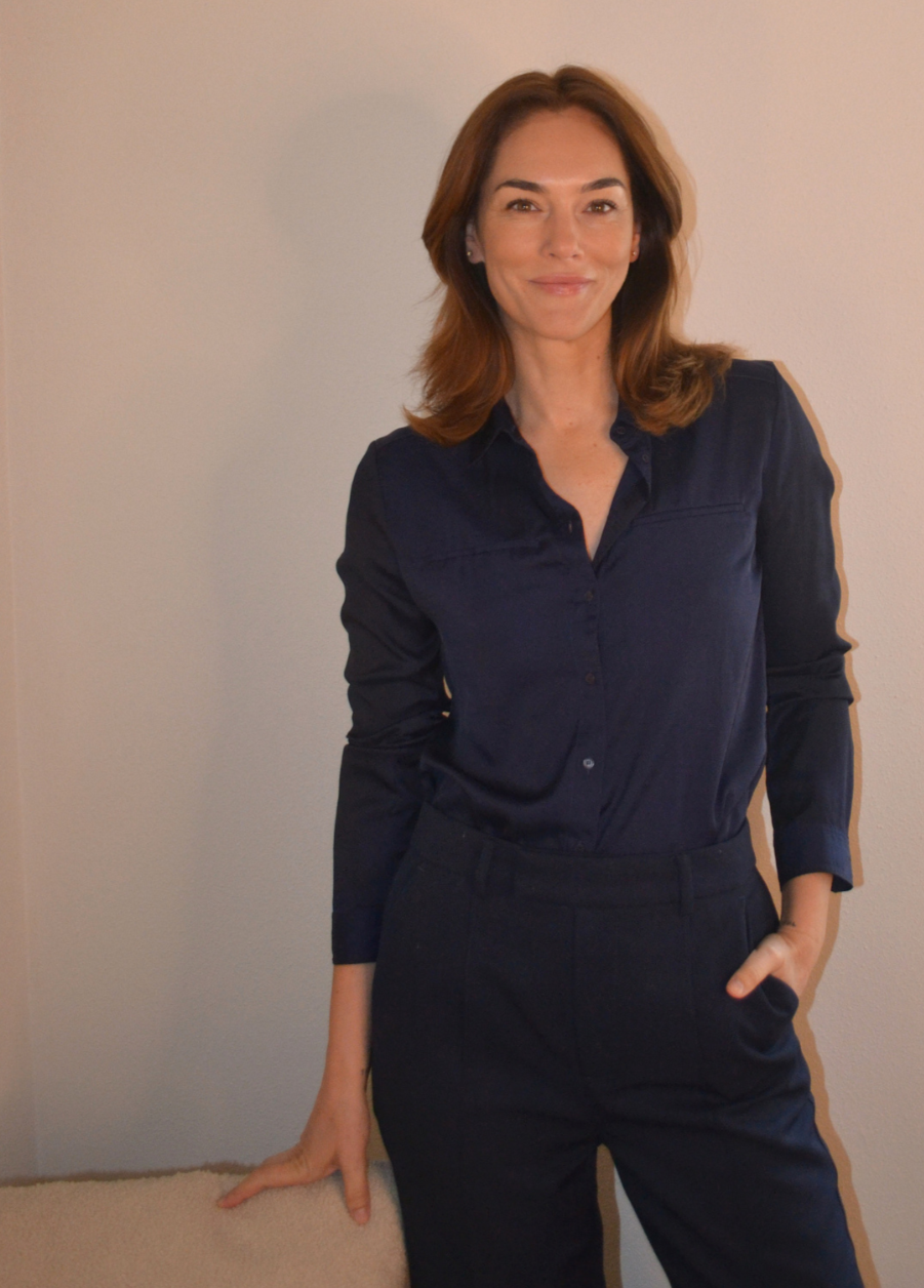 A woman with shoulder-length brown hair, wearing a navy blue button-up shirt and navy pants, standing in front of a light-colored wall, smiling.