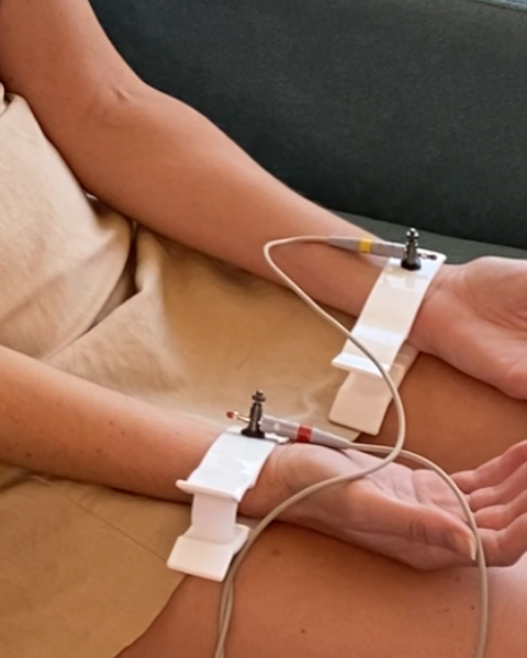 Two hands connected to medical monitoring devices with wires and electrodes, resting on a person's lap.