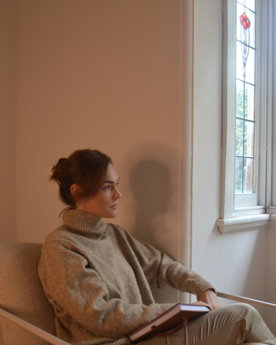 Woman sitting on a beige chair with a book on her lap, near a window with stained glass, casting a shadow on the wall.
