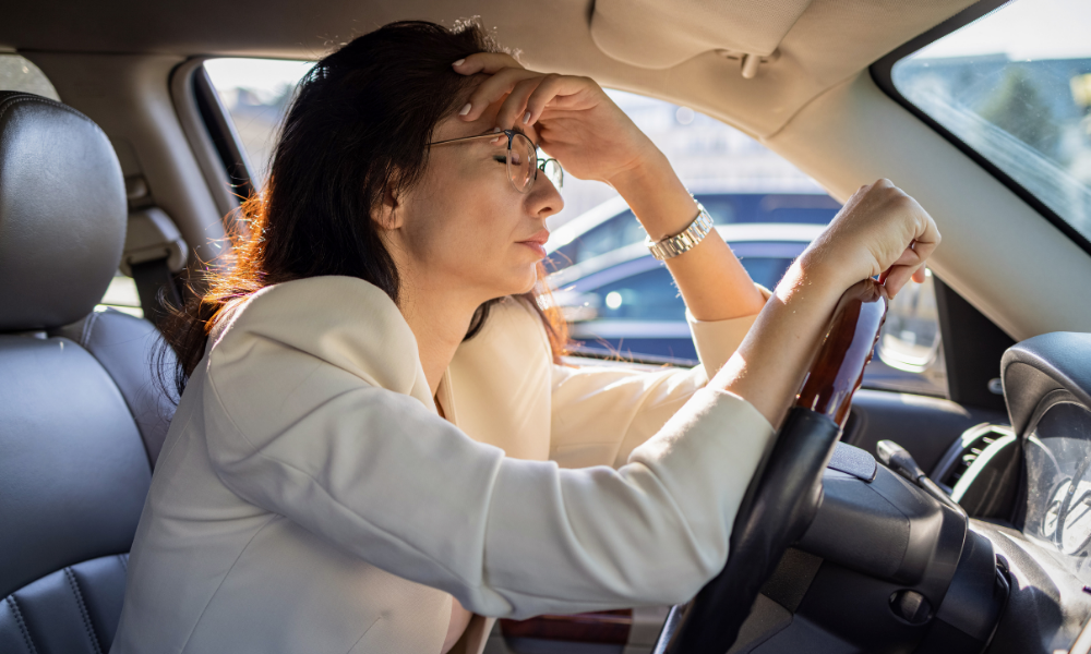 A woman sitting in the driver's seat of a car with her head resting on her hand, appearing stressed or exhausted.