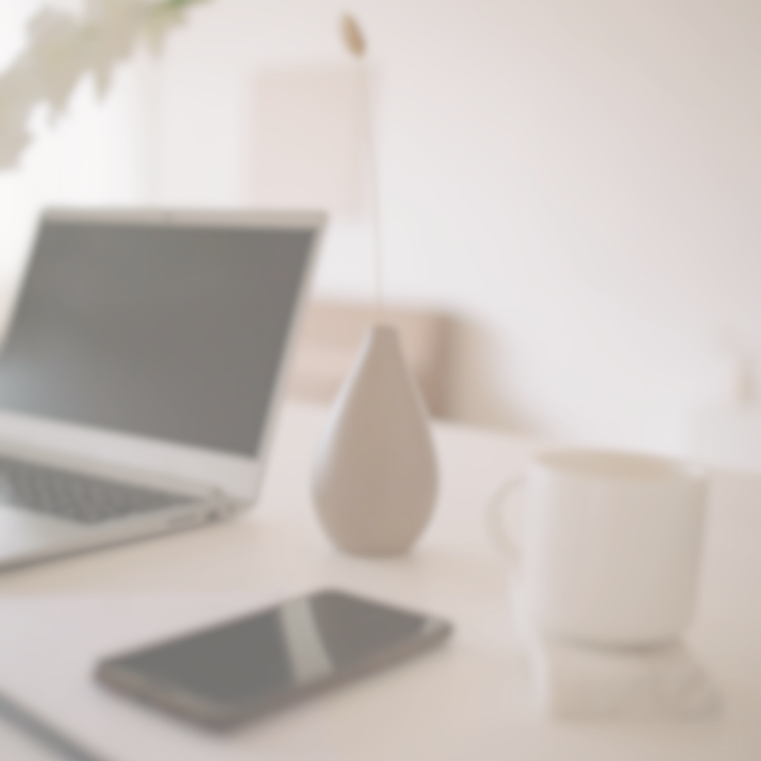 Blurry image of a white desk with a laptop, a smartphone, a white vase, and a white mug, with a plant in the background.