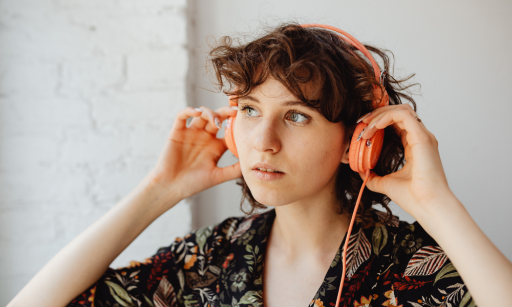 A woman with curly brown hair wearing orange headphones, adjusting them on her ears, and looking off to the side.