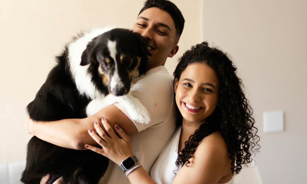 A smiling young woman with curly hair holding a happy man and a black and white dog.