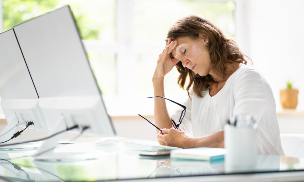 Stressed woman holding her head and glasses at her desk with a computer and notebooks.