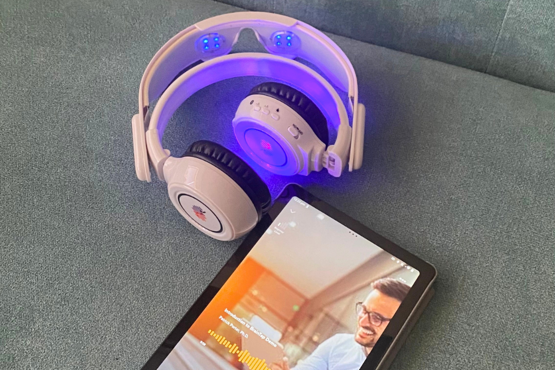 A pair of white wireless headphones with blue LEDs resting on a gray surface next to a tablet displaying a person on a video call.