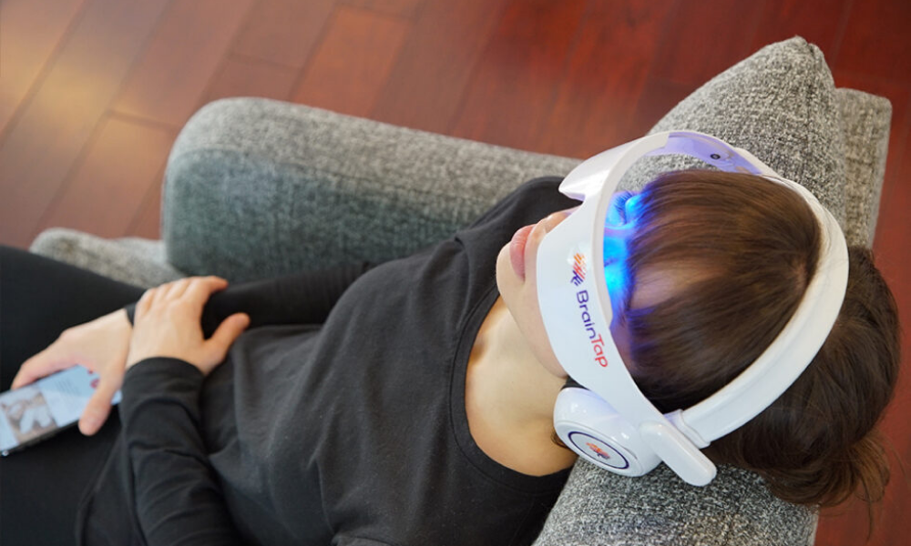 Person lying on a couch with a headband and headset labeled "Brain TEP", using a smartphone.