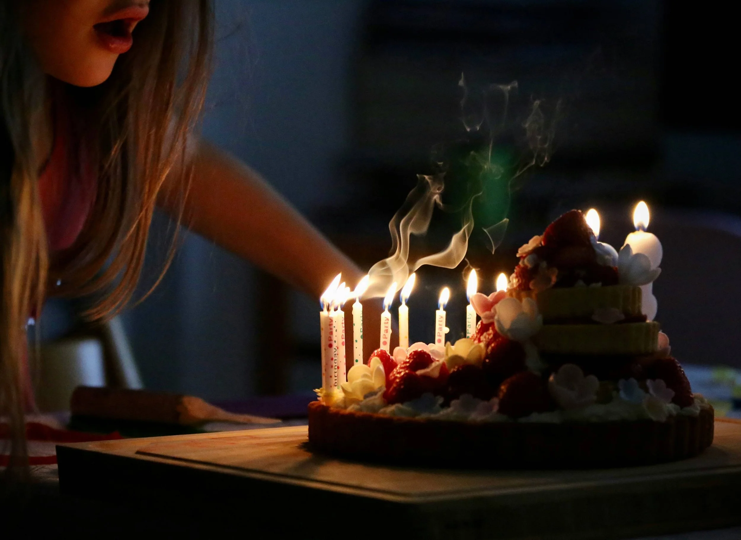 Close-up of a girl blowing out birthday candles on a decorated cake with strawberries, flowers, and cream, in a dark setting.